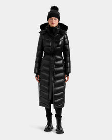 A person stands against a plain light background wearing the Rudsak ELSIE S - L124536 Black women’s shiny black, water-resistant maxi down puffer with fur-trimmed hood, paired with a black beanie with pom-pom, black boots, and pants.