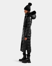 A woman in profile wears the Rudsak ELSIE S - L124536 Black maxi down puffer with a fur-lined hood, black winter boots, and a pom-pom beanie against a plain white background. The coat is insulated with RDS-certified down for warmth.