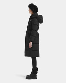 In profile, a person stands against a white background wearing the Rudsak EMBER M-L - L125543 Black women's long down puffer coat with removable hood, paired with a black fur hat, black leggings, and black lace-up boots.