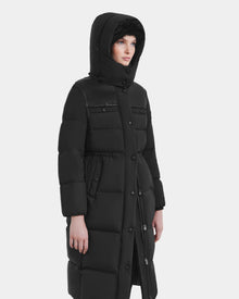 A person wearing the Rudsak EMBER M-L - L125543 Black women's long down puffer coat with a removable hood, visible buttons, and two chest pockets stands slightly to the side against a plain white background.