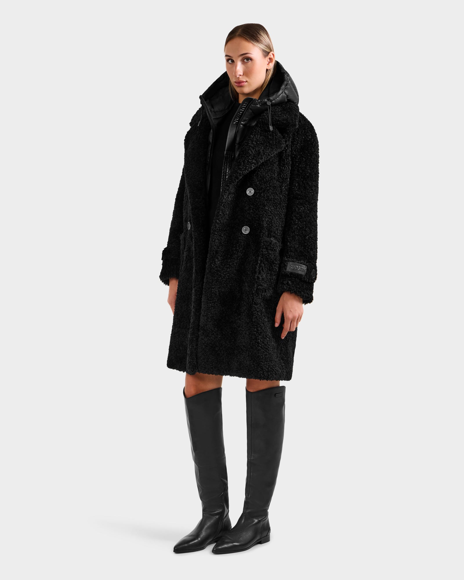 A person stands facing forward in the Rudsak EMRYN - L125588 Black textured winter coat over a hooded layer with knee-high black boots, set against a plain light gray background.