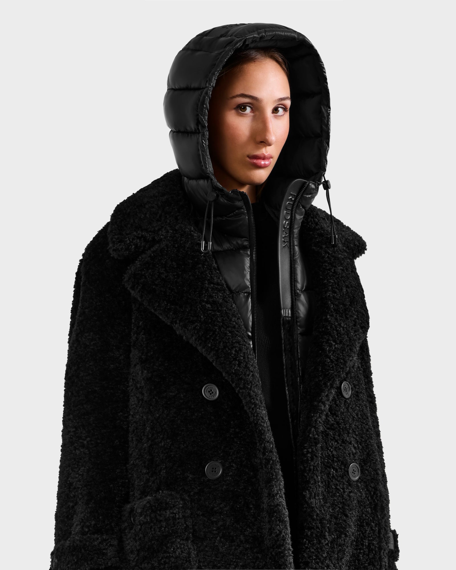 A person in the Rudsak EMRYN - L125588 Black textured winter coat layered over a shiny black hooded jacket gazes ahead with a neutral expression against a plain light background.