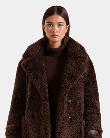 A woman with straight brown hair wears Rudsak’s EMRYN - L125588 Espresso teddy jacket over a brown turtleneck, standing against a plain, light background.