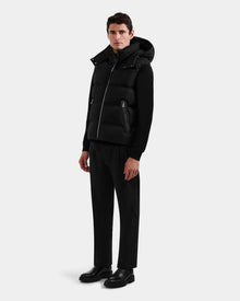 A man stands at an angle against a light gray background, wearing the Rudsak HARLOW - M125528 Black men’s puffer vest with a hood over a black sweater, paired with black trousers and shoes.