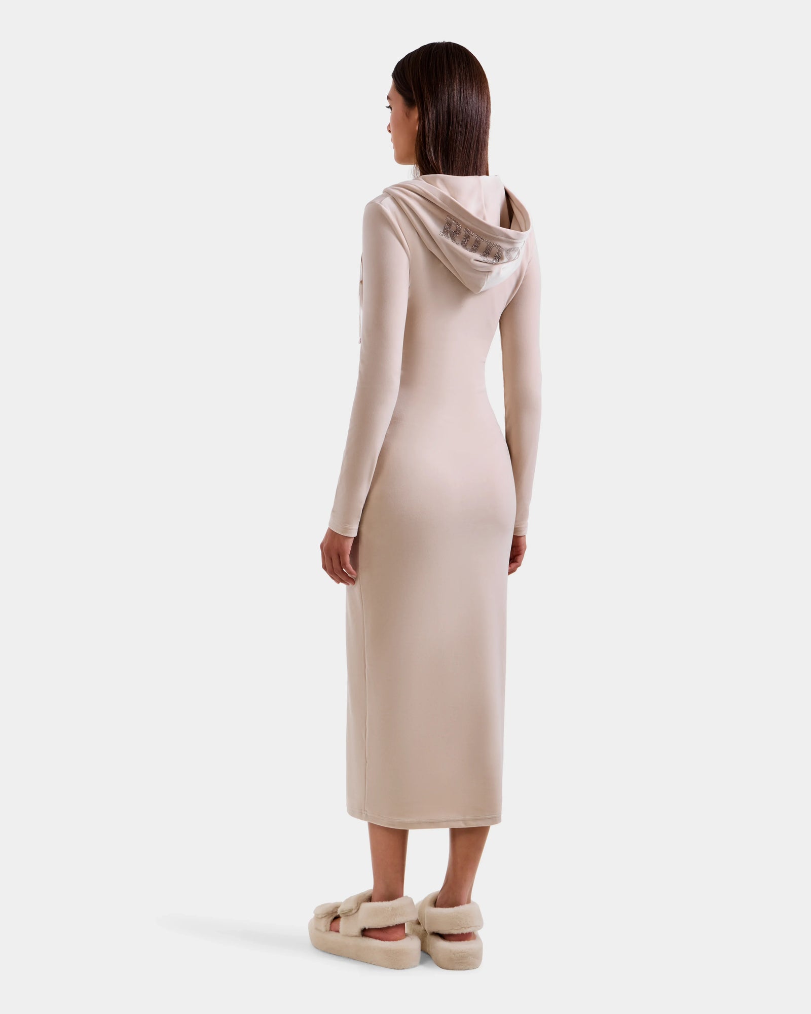 A woman faces away in the Rudsak ISABELLA - L525502 Cream, a long, fitted velour hooded dress with long sleeves and beige fuzzy slippers. The look is subtly accented by the Rudsak logo against a plain white background.
