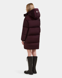 A woman with straight blonde hair, wearing the Rudsak KARLEE L - 8125682 Bordeaux long down puffer jacket with a large hood and black knee-high boots, stands facing away against a plain white background.