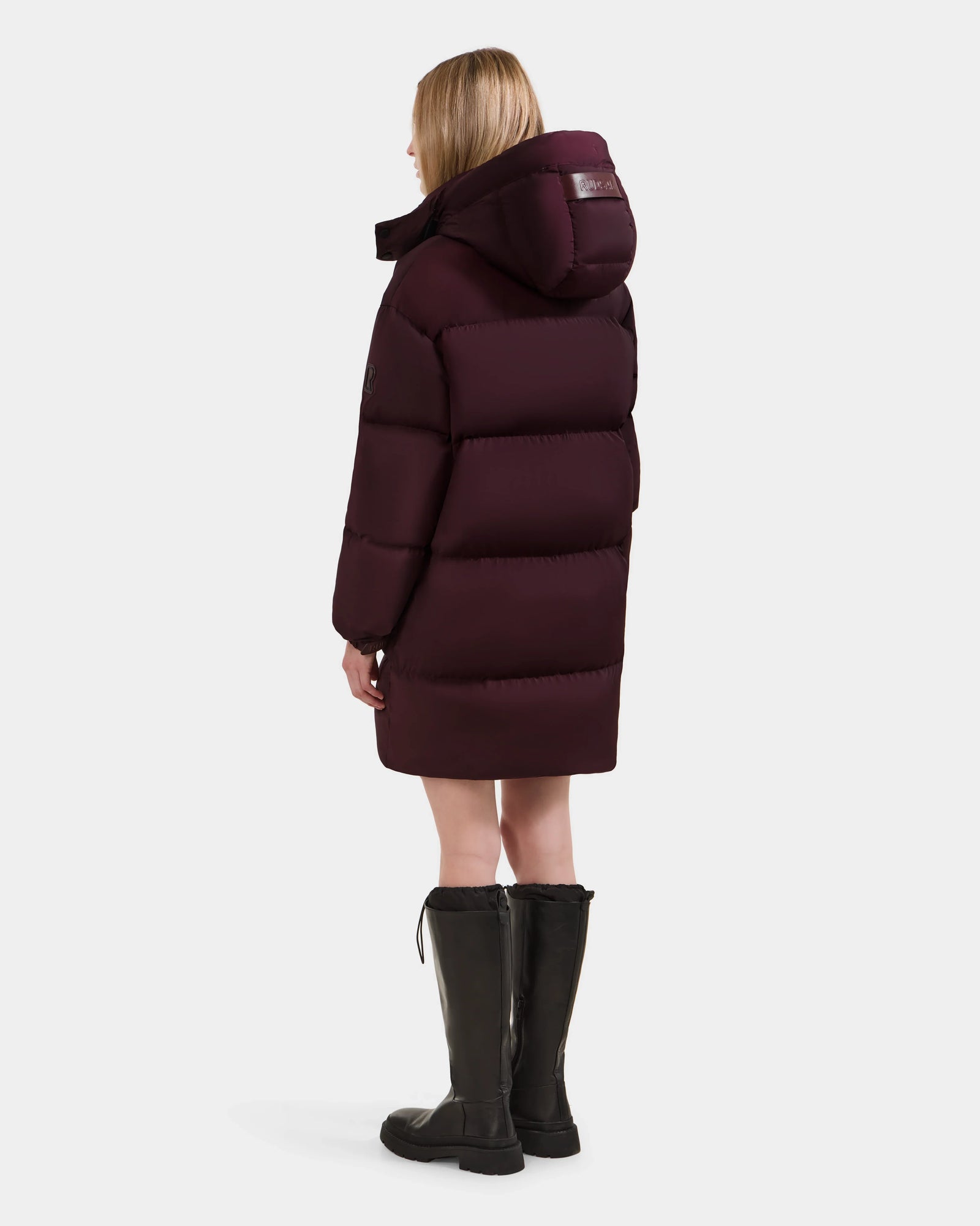 A woman with straight blonde hair, wearing the Rudsak KARLEE L - 8125682 Bordeaux long down puffer jacket with a large hood and black knee-high boots, stands facing away against a plain white background.