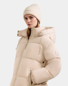 A woman wearing the Rudsak KARLEE L - 8125682 Vanilla long down puffer jacket with a hood and matching beanie stands against a plain white background, looking slightly to the side with a neutral expression.