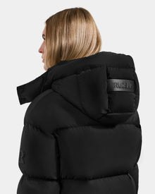 A person with straight blonde hair, shown from the back and side, wears the Rudsak KARLEE M - 8125681 Black women’s down puffer jacket with a high collar, hood, and RUDSAK patch—ideal for winter warmth.
