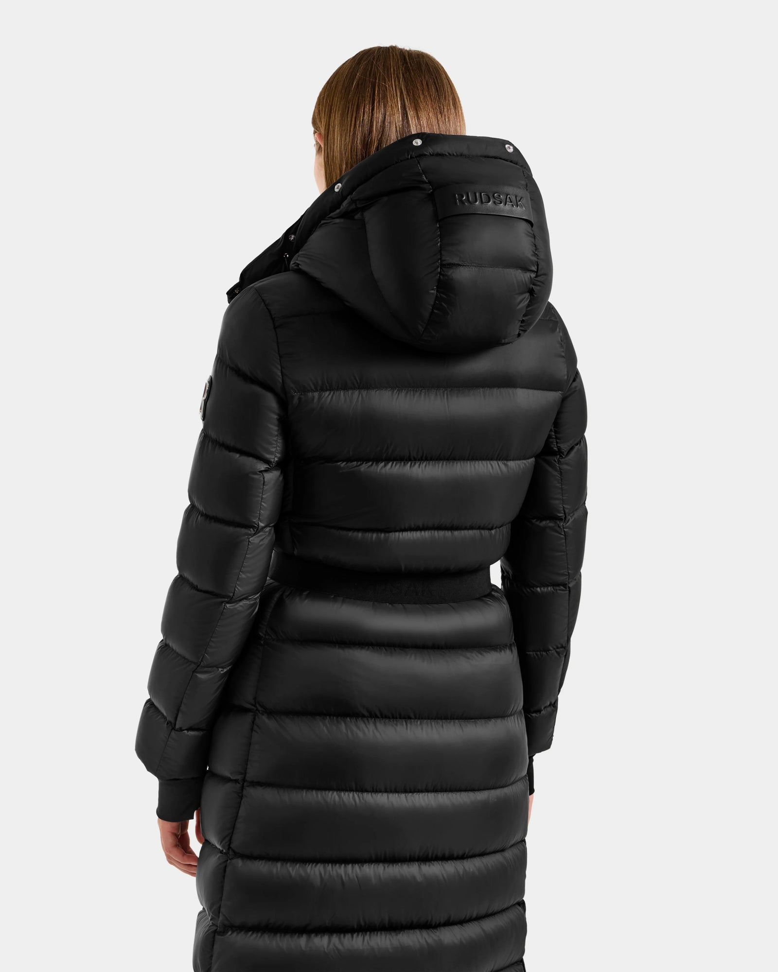 A person is seen from the back wearing the Rudsak LILITH L - L125528 Black puffer coat with a hood featuring horizontal stitching and "RUDSAK" on the hood, set against a plain, light background.