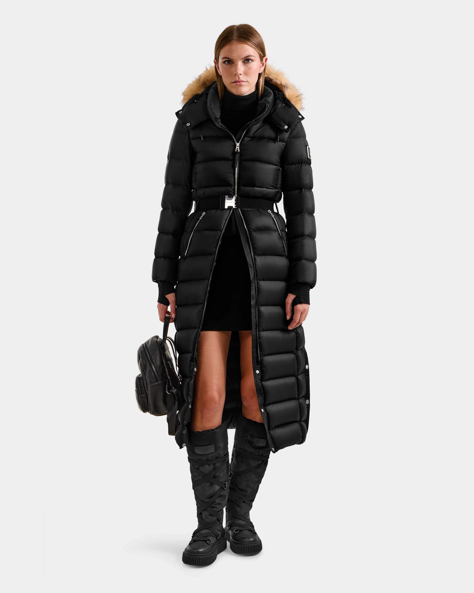 A woman faces forward wearing Rudsak's LILITH L - L125528 Black puffer coat with a fur-trimmed hood, a black turtleneck, a short black skirt, quilted knee-high boots, and holding a black backpack.