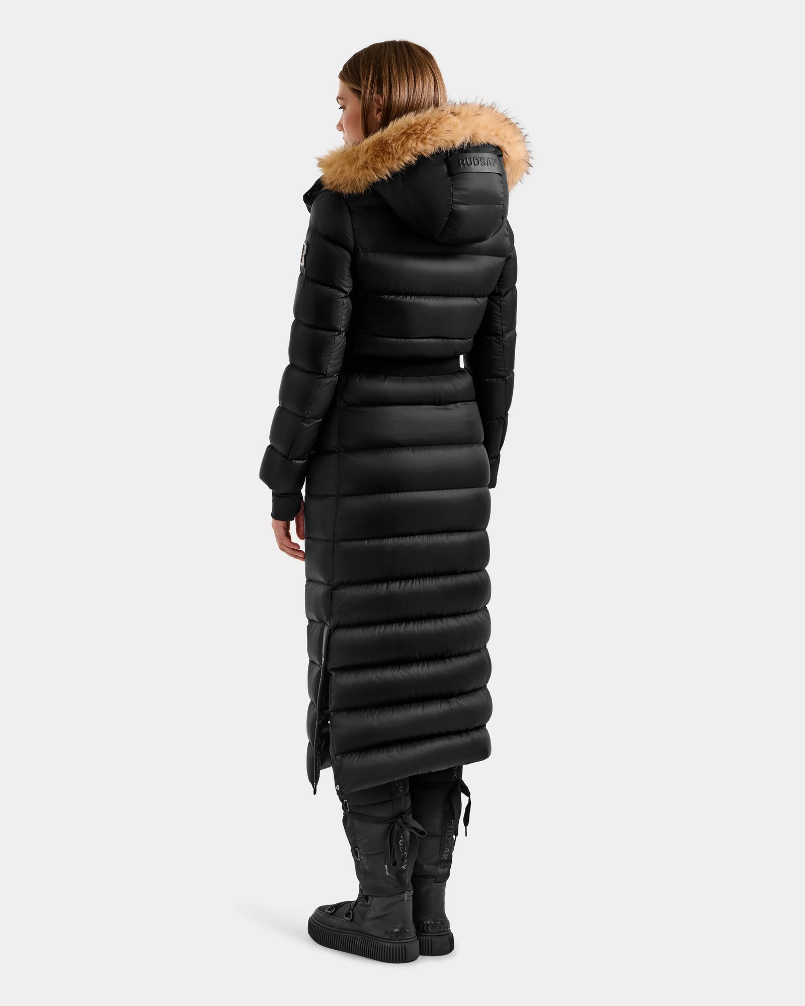 A woman stands facing away from the camera against a plain light gray background, wearing the Rudsak LILITH L - L125528 Black puffer coat with a tan fur-lined hood, paired with black pants and black boots.