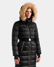 A woman wears the Rudsak LILITH L - L125528 Black maxi down puffer coat with a faux-fur trimmed hood, silver zippers, belted waist, and thumbhole cuffs, standing against a plain light gray background.