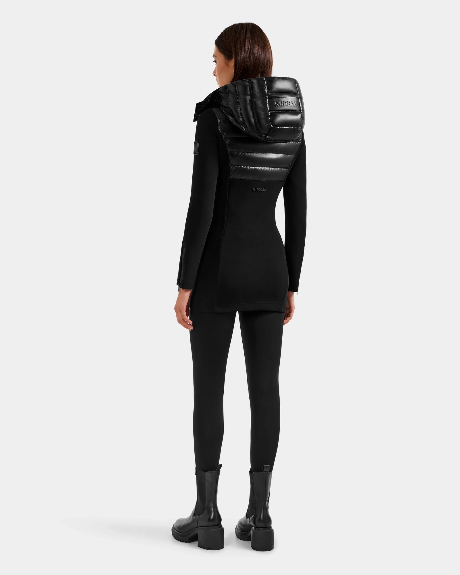 A woman faces away, wearing the Rudsak MARRY S - L125521 Black glossy shell jacket with a shiny padded hood, paired with black leggings and chunky weather-resistant boots against a plain light gray background.