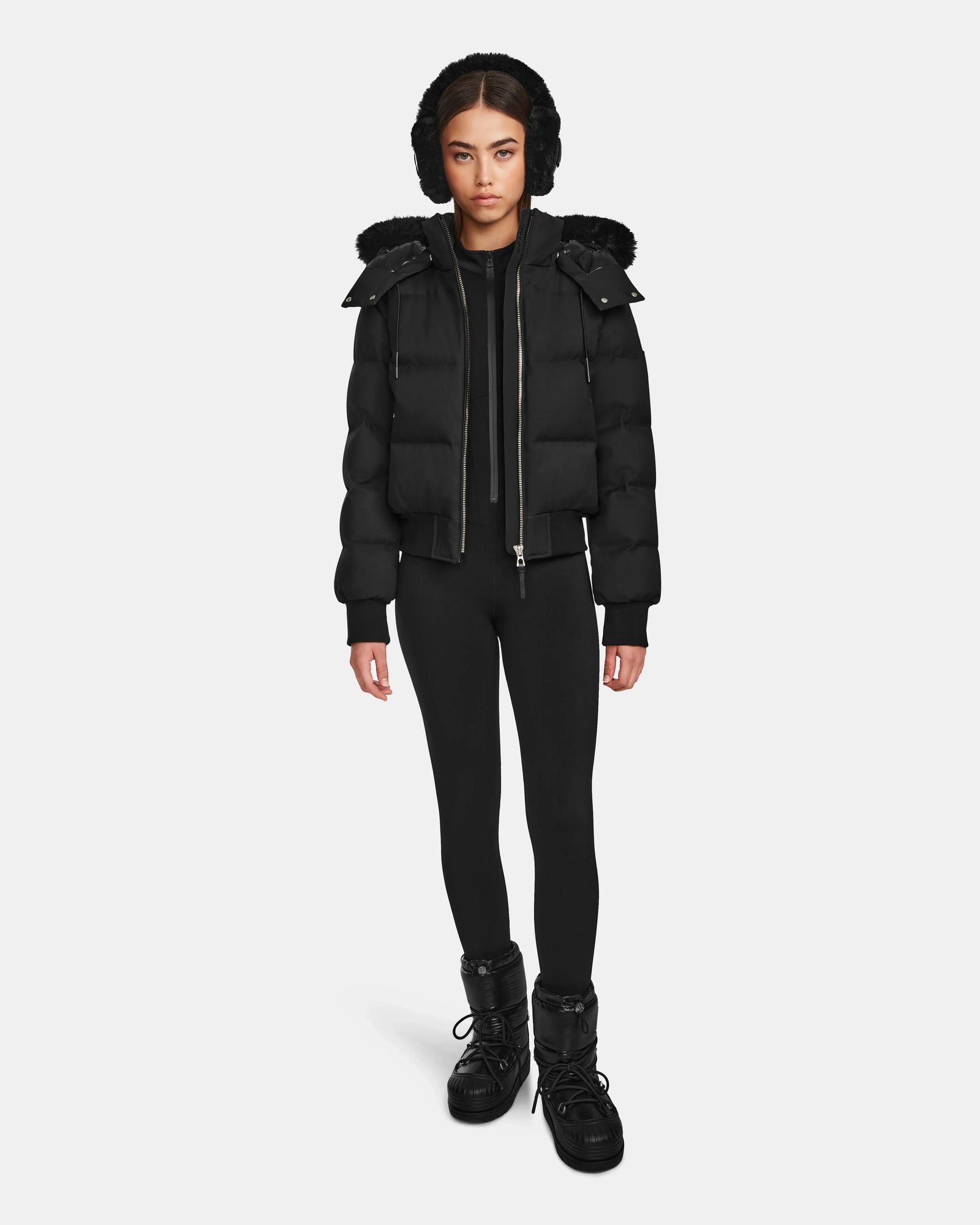 A woman faces forward, wearing the Rudsak MAUDE Q-M - L124516 Black quilted down bomber with a hood, paired with black leggings and winter boots against a plain white background.