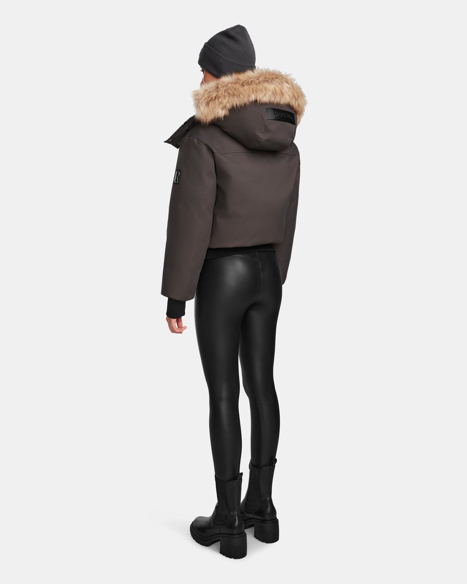 Person standing with their back to the camera, wearing the Rudsak MAUDE - L124522 Grey winter jacket with a fur-lined hood, paired with black faux-leather leggings, black boots, and a dark beanie, set against a plain white background.