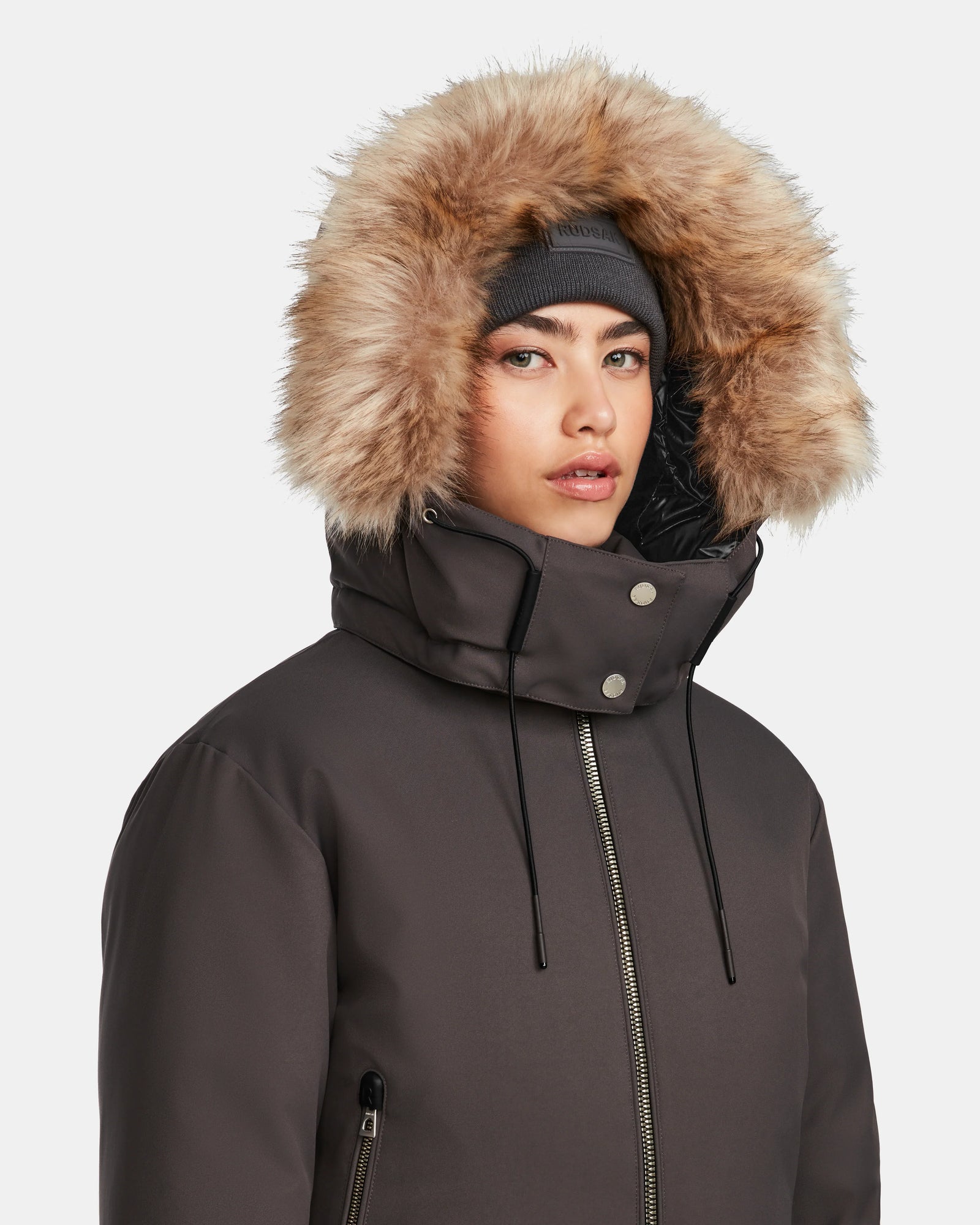 A woman models the Rudsak MAUDE - L124522 Grey down bomber, zipped up with silver snaps and drawstrings. The classic winter jacket features a fur-lined hood, premium down insulation, and is paired with a gray beanie.