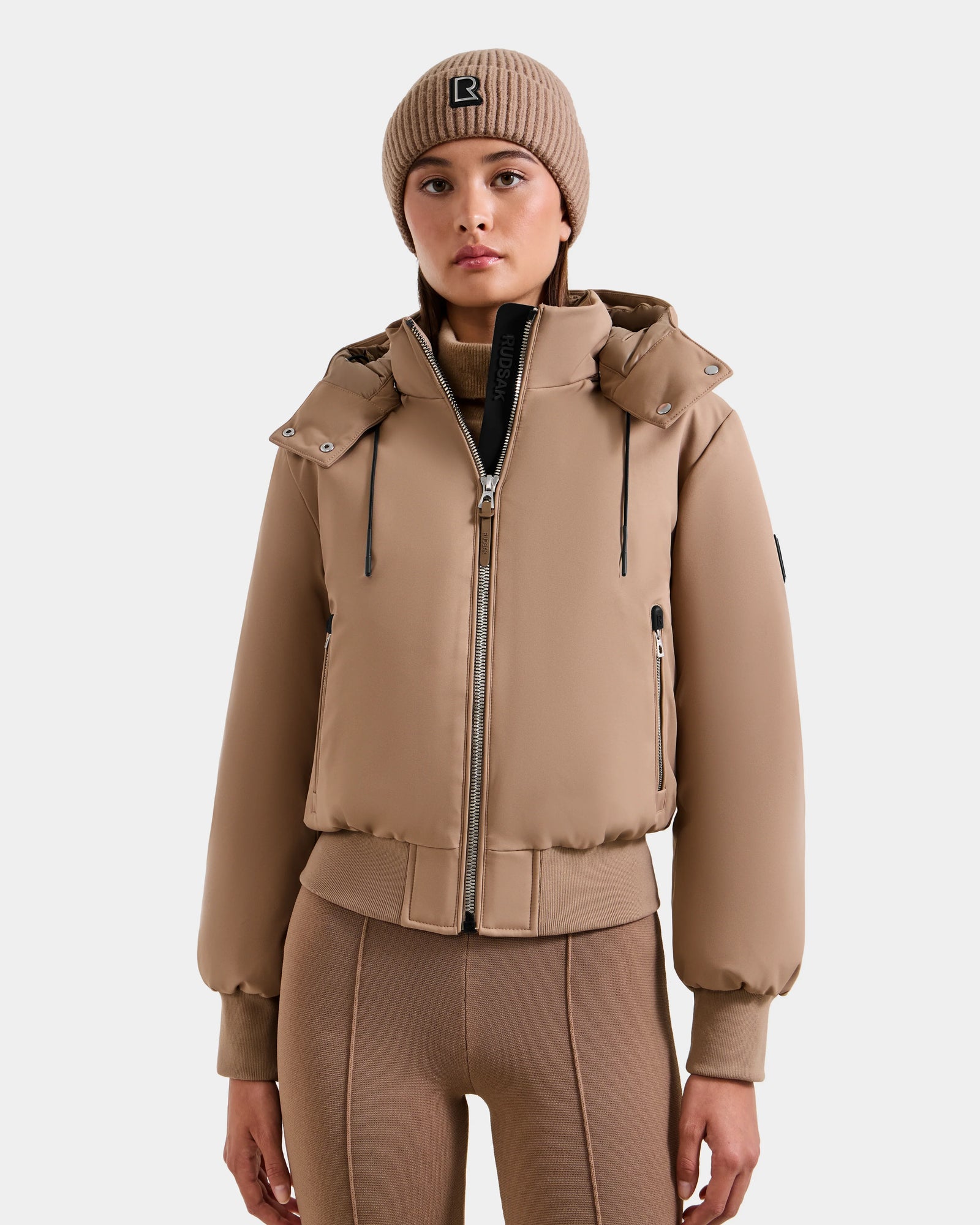 A woman in the Rudsak MAUDE - L124522 Sand down bomber, a matching beige knit logo beanie, and fitted tan pants stands facing forward against a plain white background.