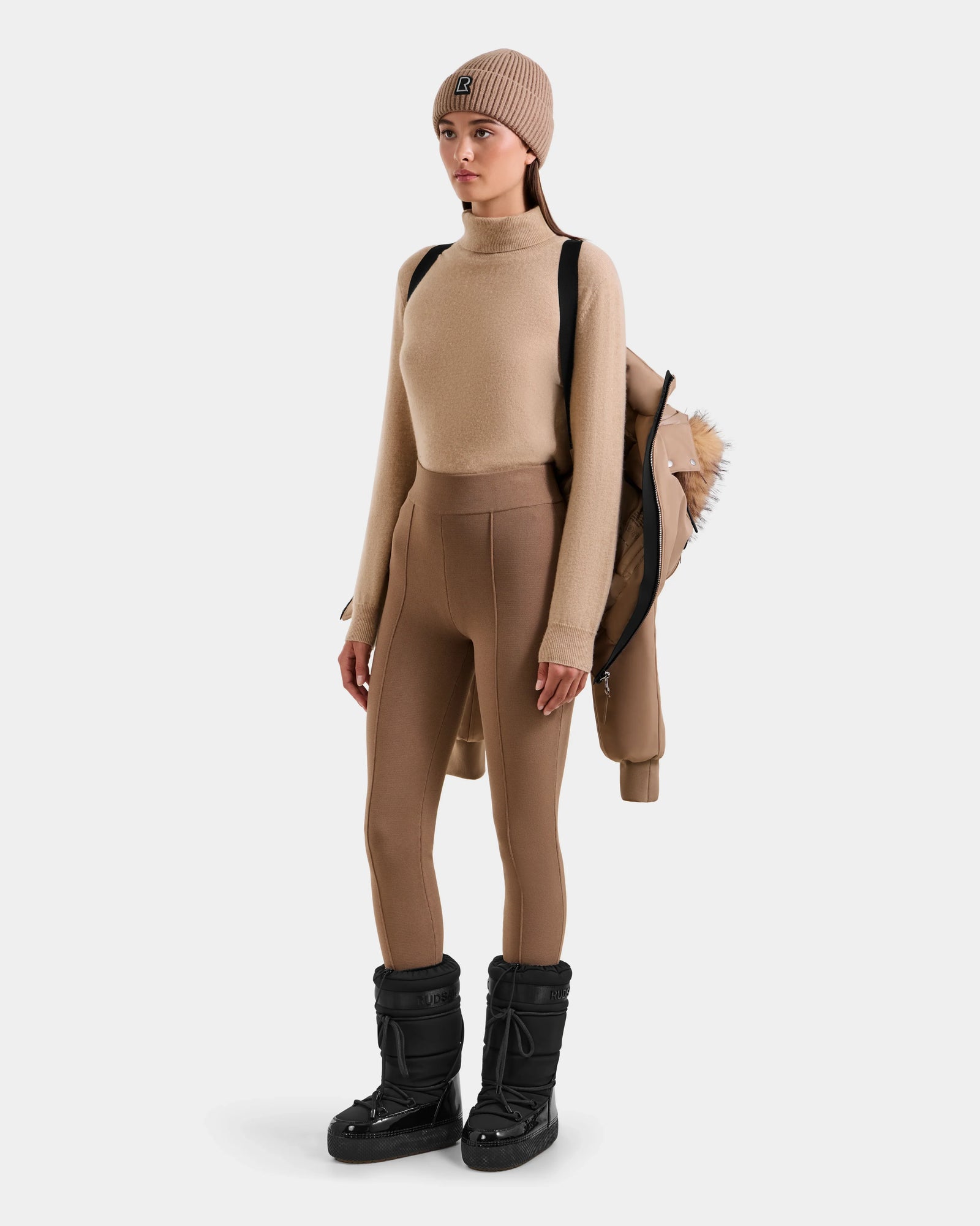 A person faces forward against a plain background, carrying the Rudsak MAUDE - L124522 Sand tan winter jacket with fur trim and premium down insulation, while wearing a beige beanie, tan sweater, fitted pants, and black boots.