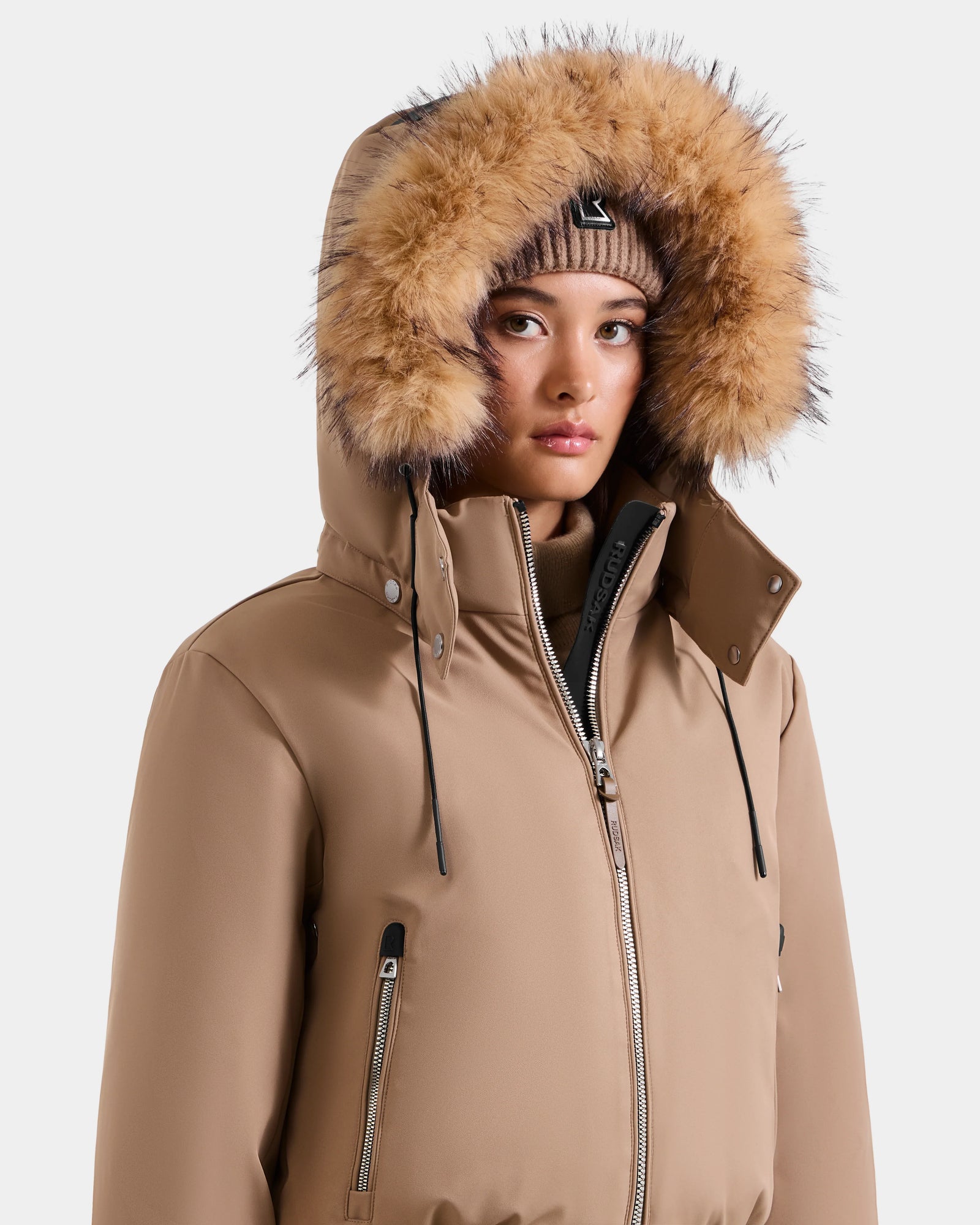 Wearing the MAUDE - L124522 Sand women’s down bomber by Rudsak, featuring premium down insulation and a fur-lined hood, paired with a brown knit beanie. The zipped-up jacket’s hood frames her face against a plain light background.