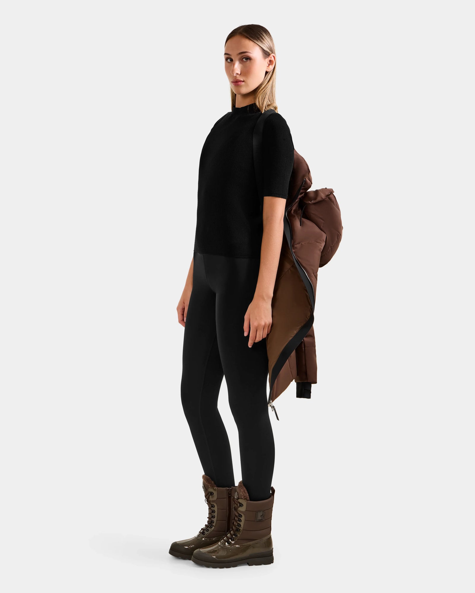 A woman stands sideways against a white background, wearing Rudsak's MIRIAM - L123544 Mocha women's down puffer jacket with a weather-resistant shell draped over one shoulder, paired with black leggings, a black top, and brown winter boots.