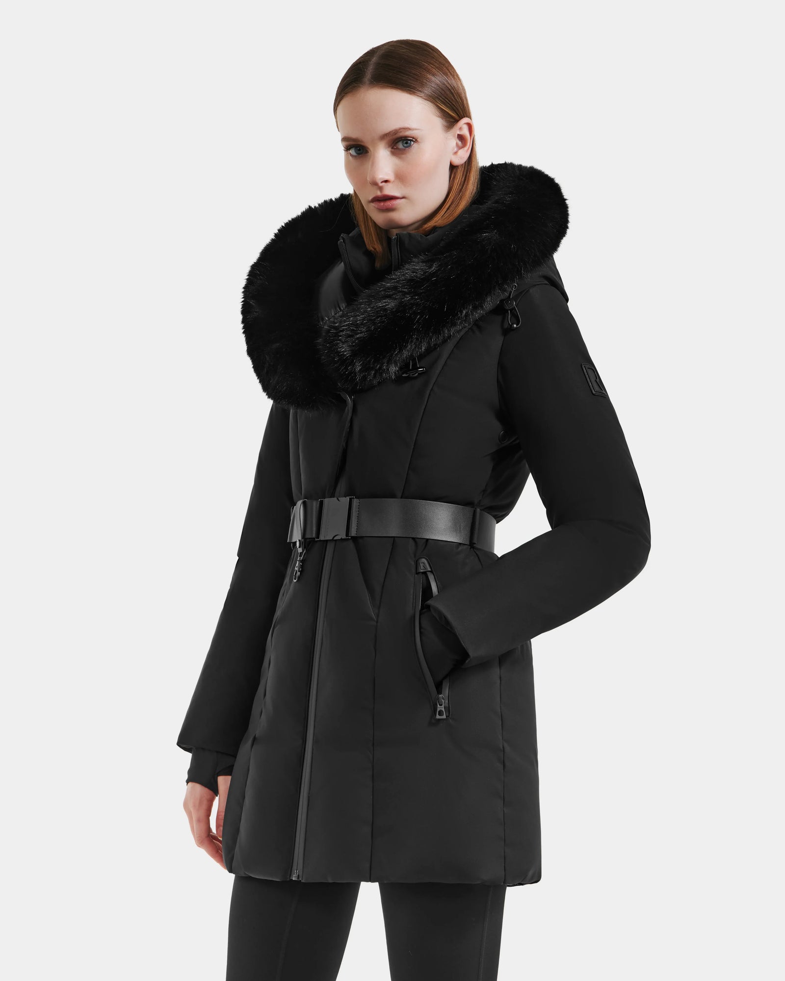 A woman with straight brown hair and a neutral expression models the Rudsak MODA M - L125555 Black long belted winter coat with a large faux fur collar against a light gray background, her left hand in the pocket—ideal for winter layering.