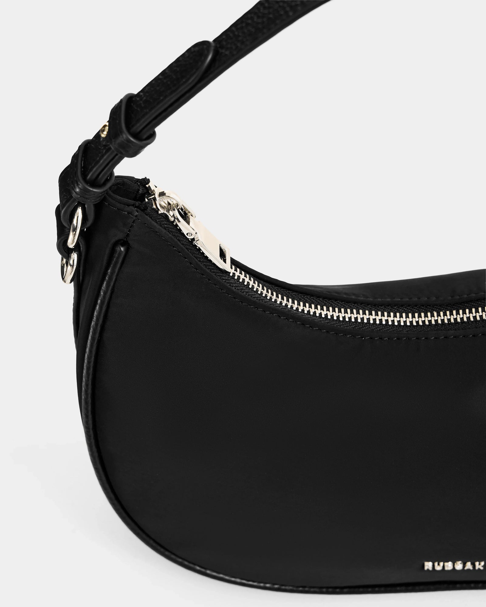 Close-up of the Rudsak NIKKI - L325512 Black women's crescent shoulder bag featuring a gold zipper, adjustable strap, and gold logo on the lower right corner, set against a plain light gray background.