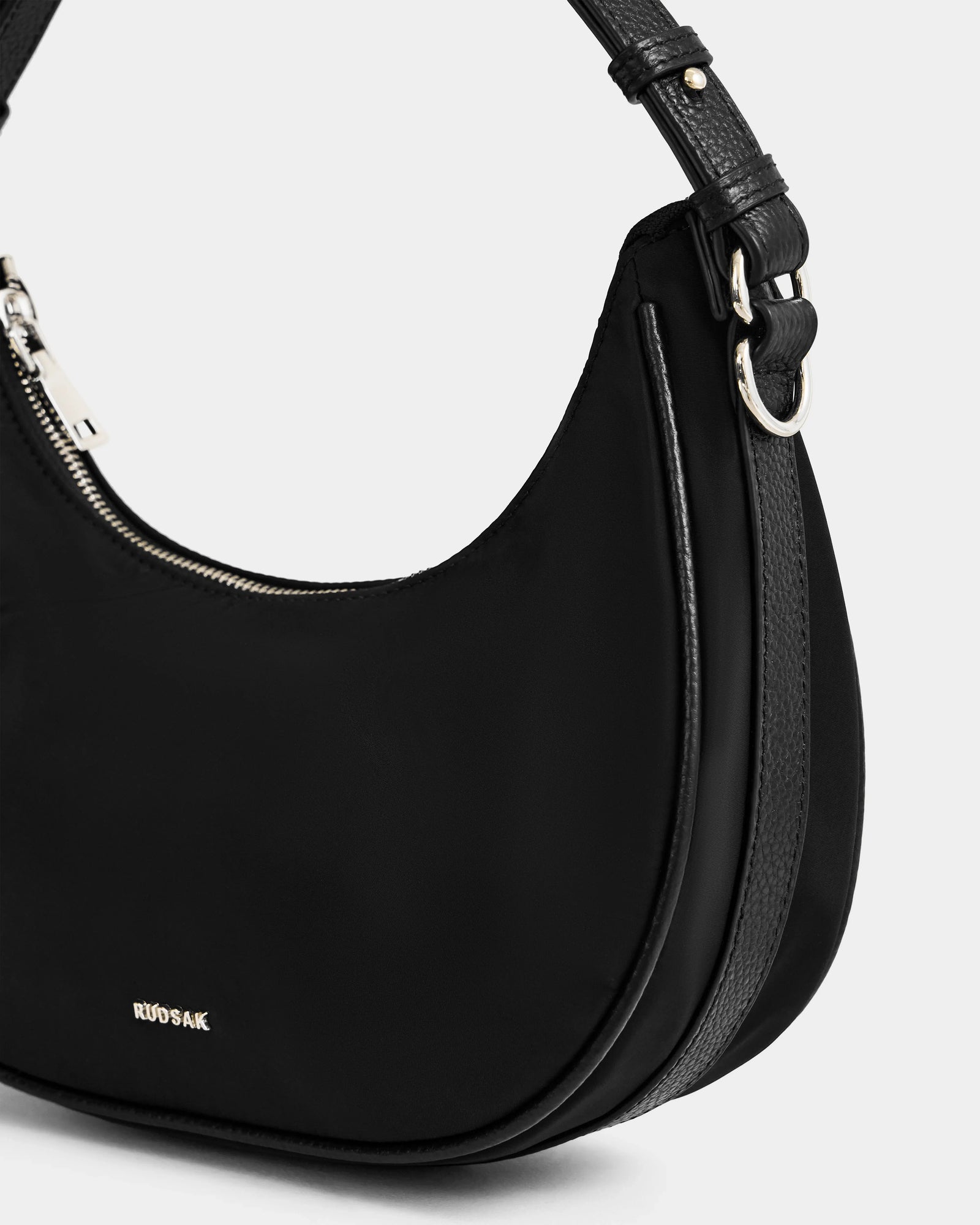 Close-up of the NIKKI - L325512 Black crescent shoulder bag by Rudsak, crafted in lightweight technical fabric with a curved design, silver zipper, textured strap, and metallic Rudsak logo. Displayed on a plain light grey background.