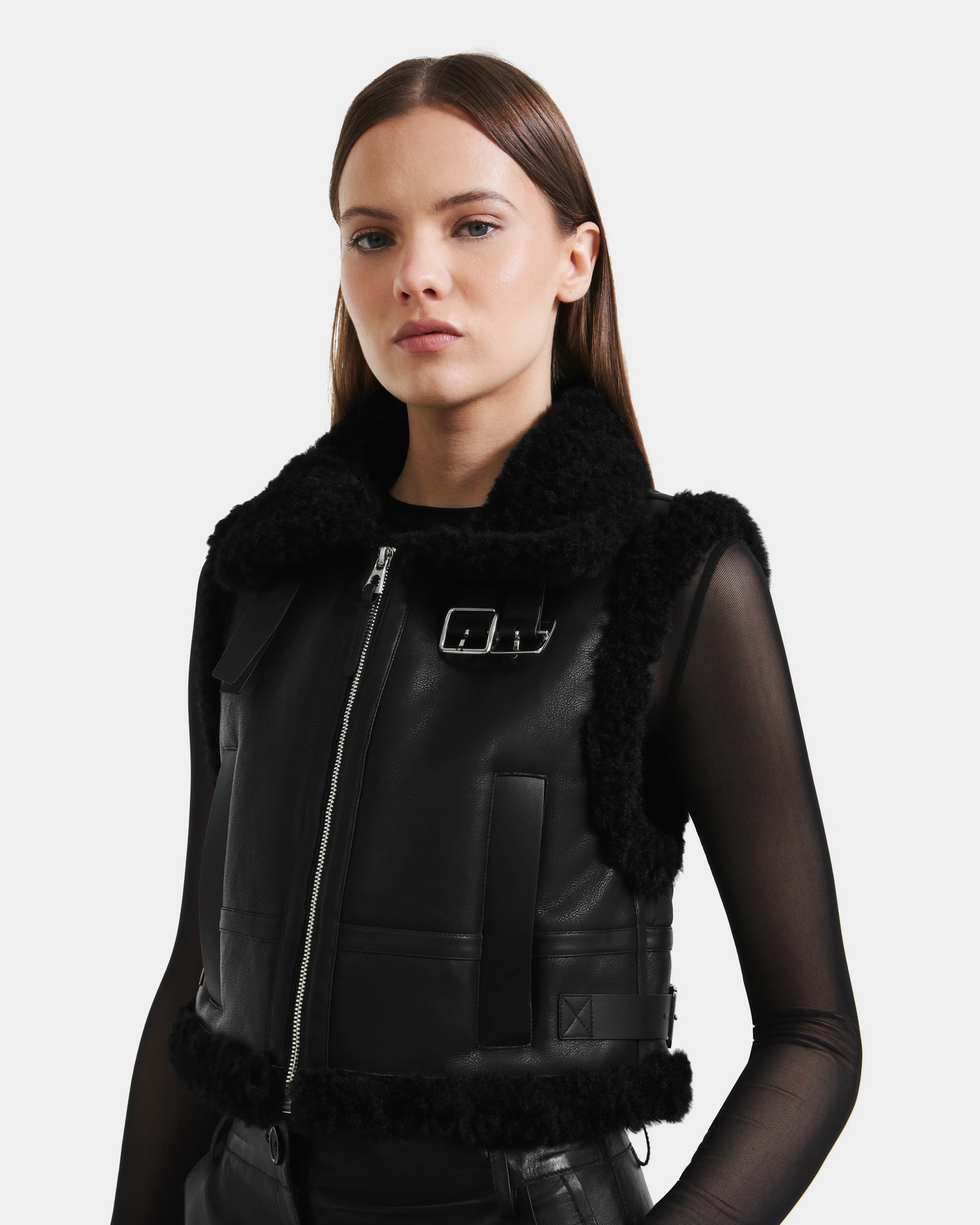 A woman with straight brown hair models the Rudsak PAISLEY - L125516 Black vegan leather cropped vest with faux shearling collar, zipper, and buckle over a sheer black top, posing against a plain light background.
