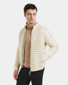 A man in a Rudsak REYMOND M - M125523 Vanilla RDS-certified down puffer jacket over a tan turtleneck and black pants stands against a white background, looking left with one hand in his pocket.