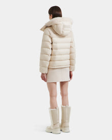 A woman faces away, modeling the Rudsak SANDRA - 8125612 Vanilla women's shearling puffer jacket with lightweight insulation, styled with a matching short skirt and fur-trimmed knee-high boots in light beige against a white background.
