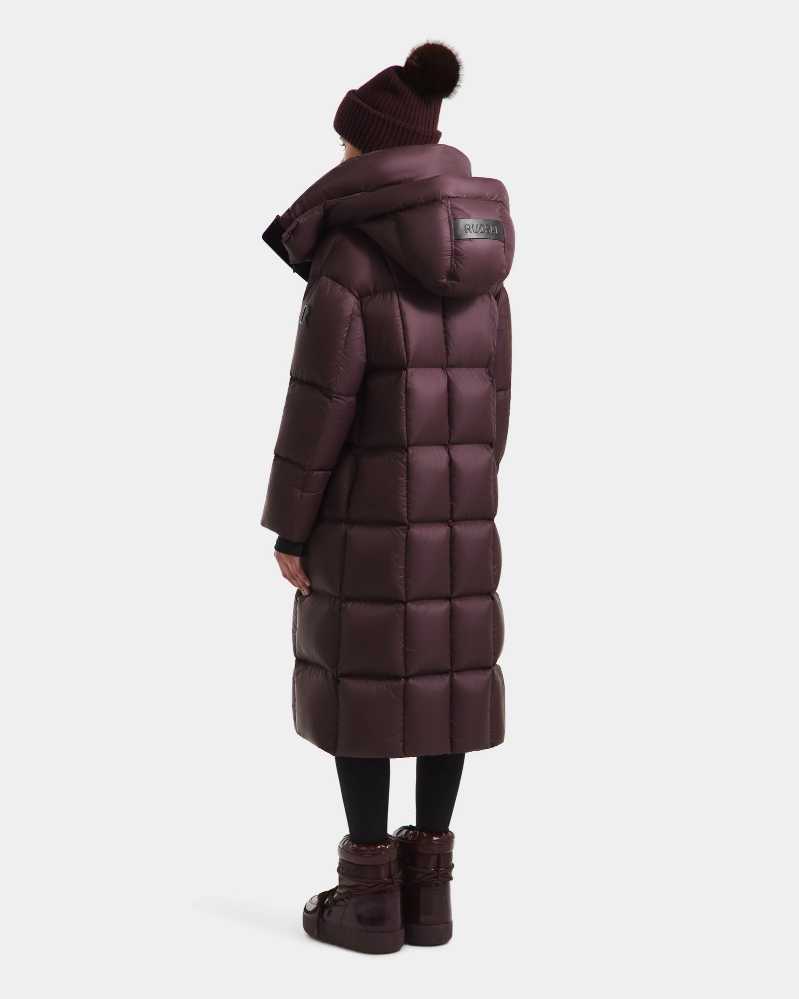 A person is seen from behind wearing the Rudsak SASHA - 8125686 Bordeaux women’s long down puffer coat with a hood, black leggings, a maroon pom-pom beanie, and matching boots.