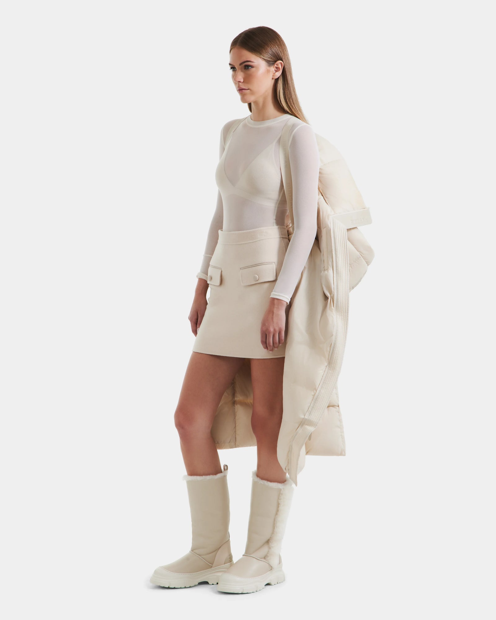 A woman in profile holds the Rudsak SASHA - 8125686 Vanilla waterproof winter coat over her arm, wearing a sheer long-sleeve top, cream mini skirt with pocket flaps, and tall cream boots against a plain white background.