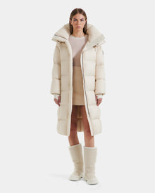 A woman faces forward in a Rudsak SASHA - 8125686 Vanilla long down puffer coat with removable hood, beige mini skirt, sheer beige top, and cream winter boots against a plain white background.