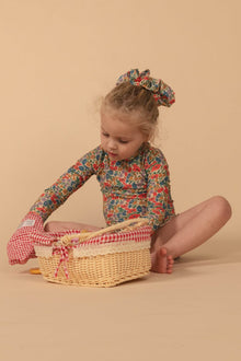 Toddler wearing long-sleeve floral rashguard in red, blue, and gold tones with matching hair accessory.