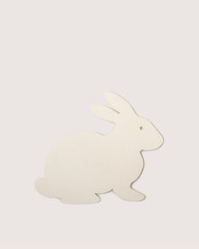 A season-exclusive Graf Lantz trivet features a simple beige flat cut-out rabbit design on a light gray background. Resembling thick paper or thin cardboard, it faces right with minimal detail, similar to Merino wool.