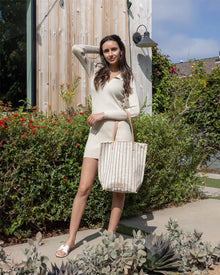 A woman in a cream long-sleeve dress stands outdoors on a path, holding the Graf Lantz Hana Canvas Tote with leather straps. She poses with one hand on her head, surrounded by greenery and flowers near a wooden wall.
