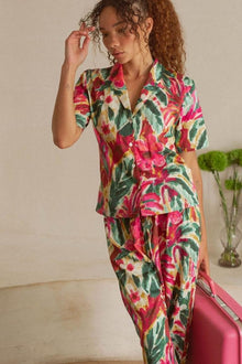Woman wearing short-sleeved floral button-up top in red, pink, yellow, and green botanical print with matching patterned pant