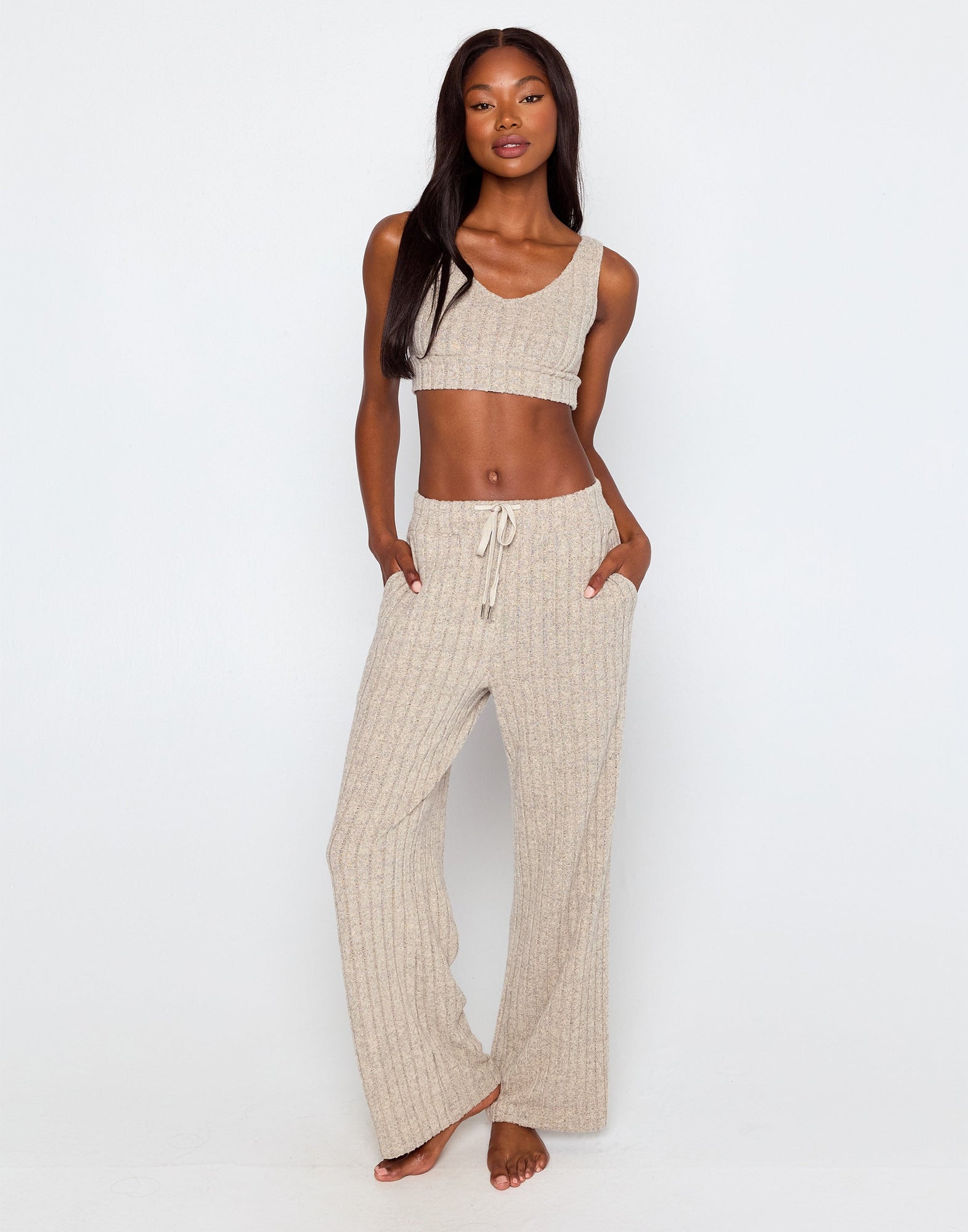 Woman wearing a beige ribbed knit loungewear set, featuring a cropped tank top with a scoop neckline and wide-leg drawstring pants with side pockets, standing barefoot against a plain white background.