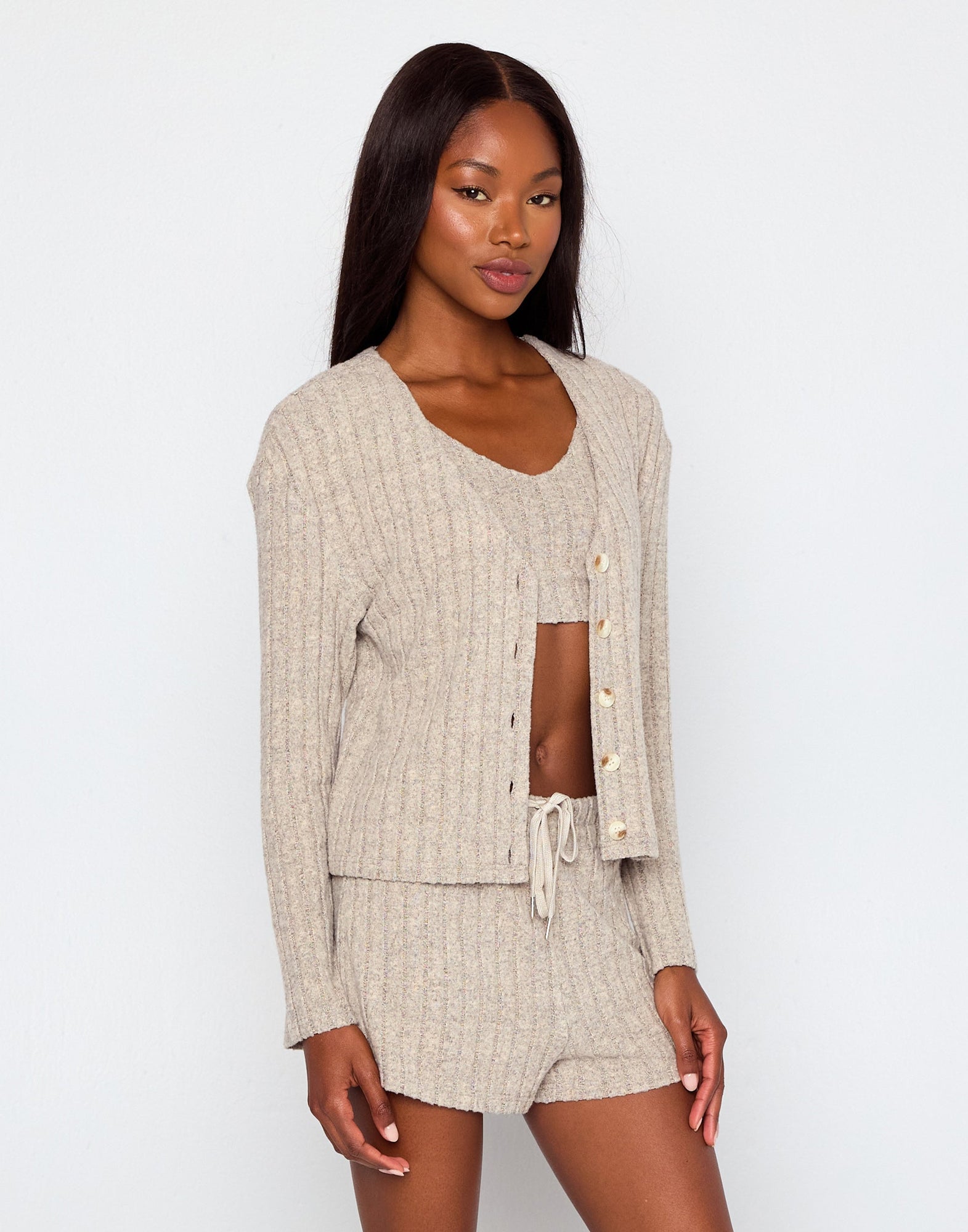 Woman wearing a beige ribbed knit loungewear set including a cropped tank top, drawstring shorts, and a matching button-up cardigan with long sleeves, posing against a plain white background.