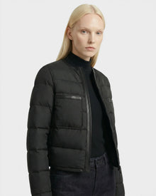 A person with straight, long blonde hair wears the Rudsak MARKLE - 8125033 Black down jacket over a black turtleneck and dark jeans, standing against a plain light gray background.
