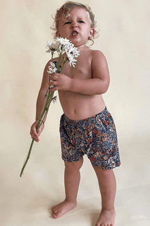 Toddler wearing navy swim trunks with multicolored floral print, holding white flowers.