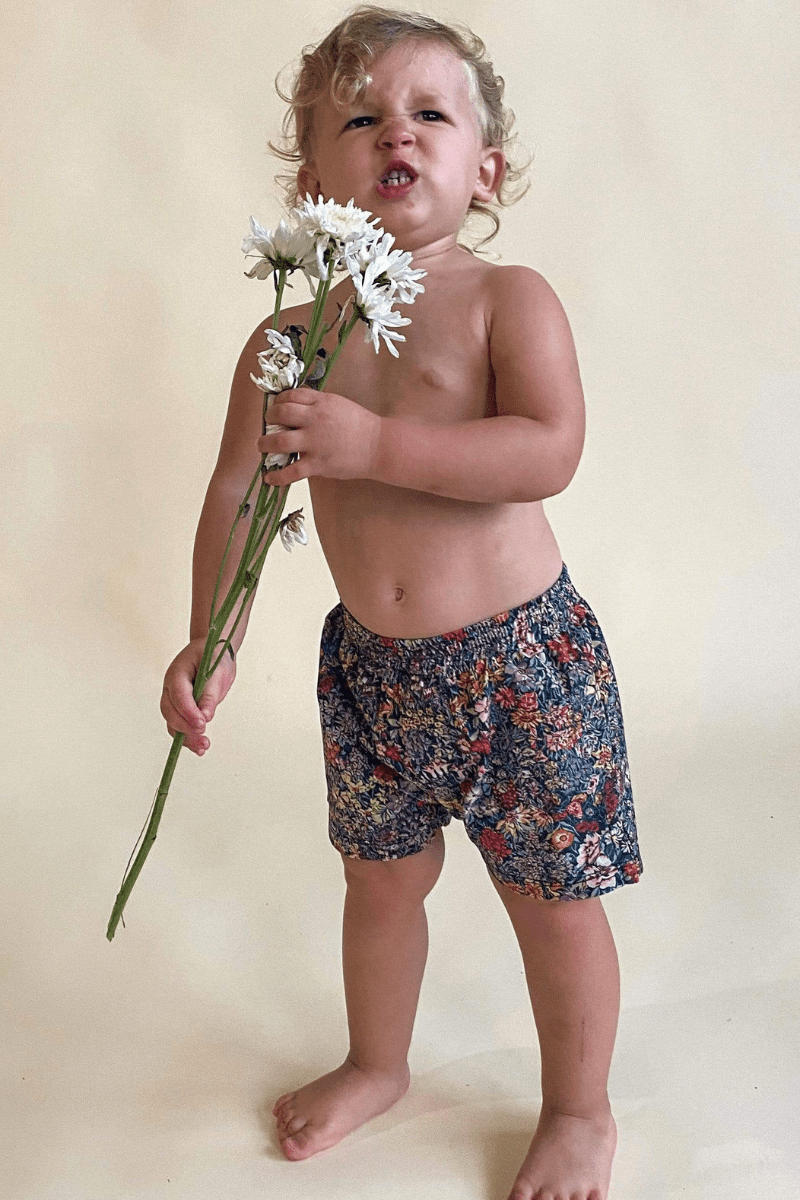 Toddler wearing navy swim trunks with multicolored floral print, holding white flowers.