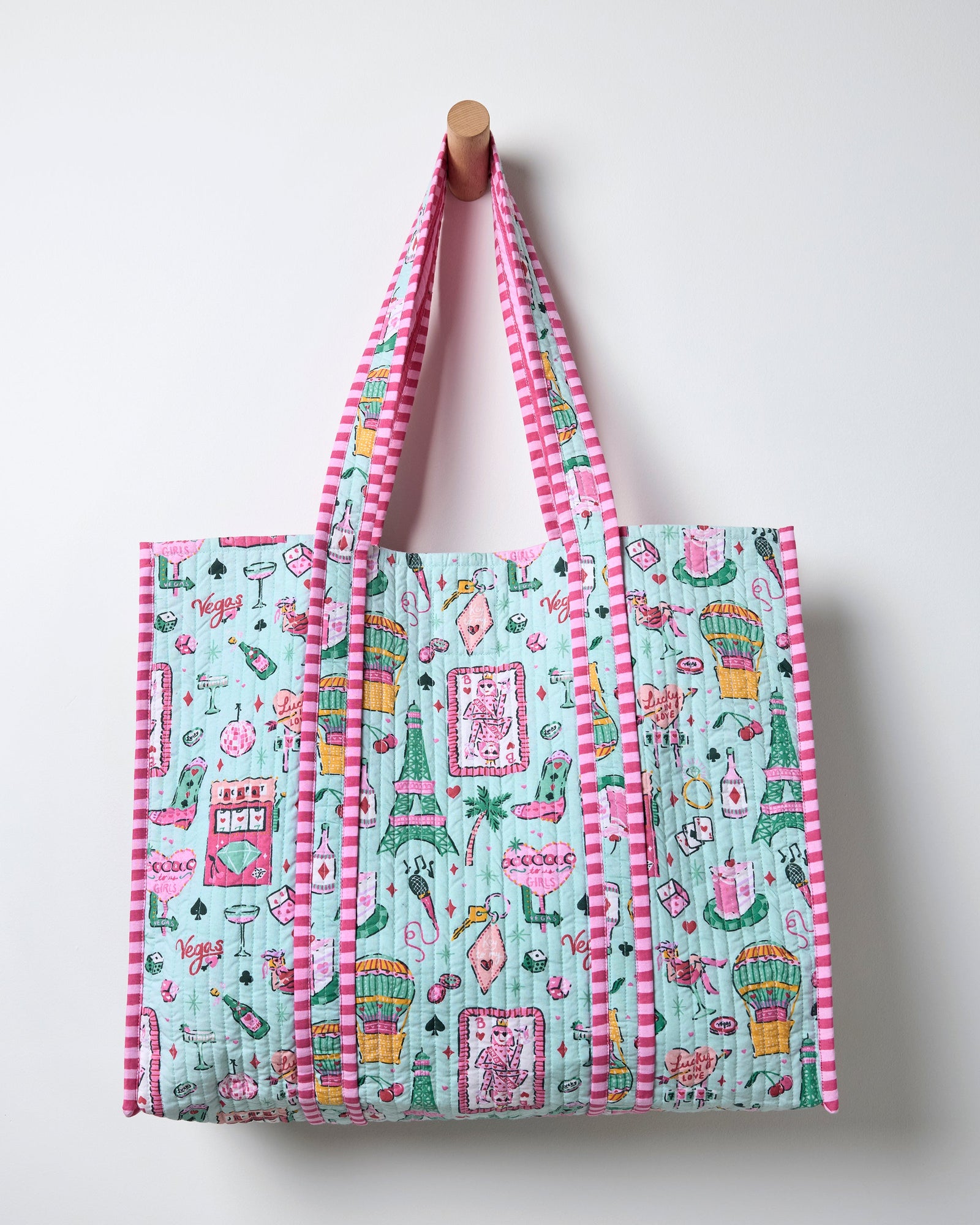 Quilted mint tote hanging from a wall peg, decorated with a colorful Las Vegas-themed print and pink striped handles and edging.