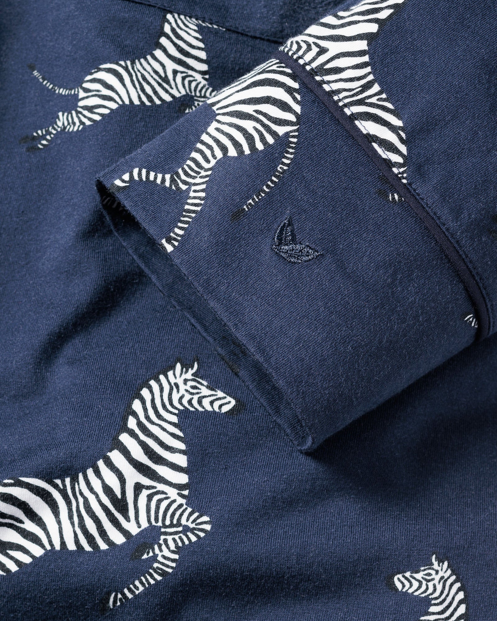 The Womens Pima Pajama Set in Zebre de Nuit by Petite Plume showcases navy blue fabric with a white and black zebra pattern. Made from premium Peruvian Pima cotton for comfort, it features a cuff adorned with a subtle sailboat logo embroidery.