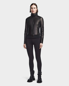 A woman stands facing forward wearing the Rudsak PRESLEY - 8122027 BLACK leather jacket, crafted from supple lamb leather, paired with black leggings and black ankle boots against a light background.
