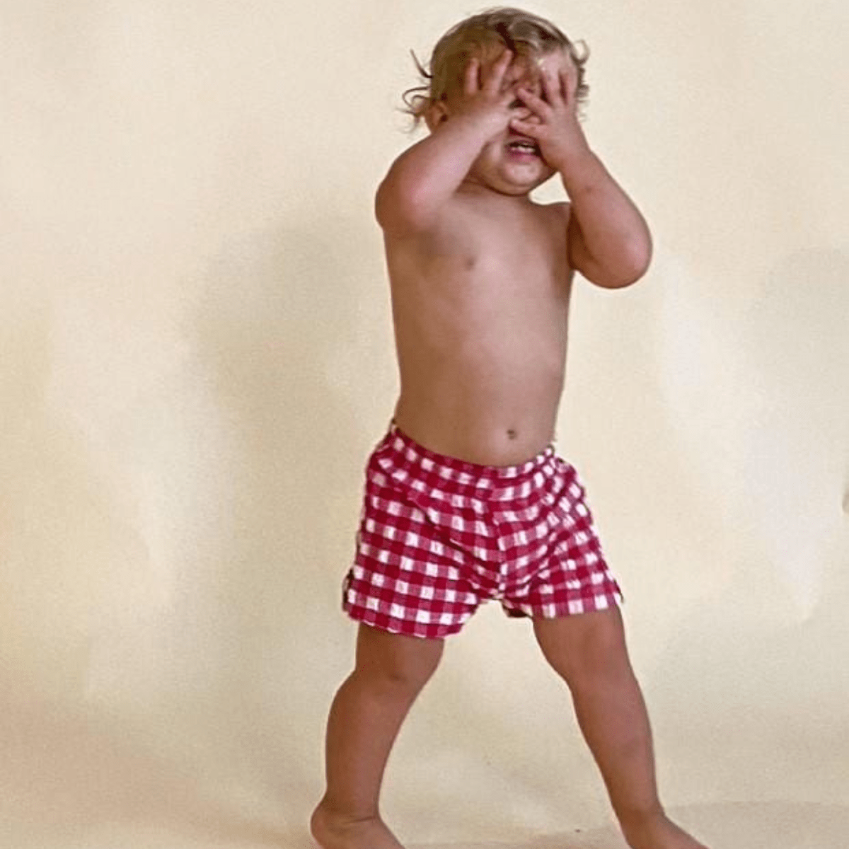 Toddler wearing red and white gingham swim trunks with elastic waistband, standing against cream background.
