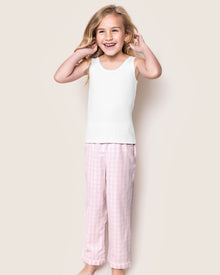 A smiling girl wearing Petite Plumes Kids Twill Pajama Pants in Pink Gingham and a white sleeveless top stands against a plain light background. Her long blonde hair cascades around her face as she gently rests her hands near her cheeks.