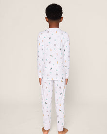 A child stands barefoot on a neutral backdrop, wearing Petite Plumes luxurious Kids Pima Snug Fit Pajama Set in Jingle Paws. The set features soft Pima cotton with colorful cartoon animal prints and long sleeves for added comfort.