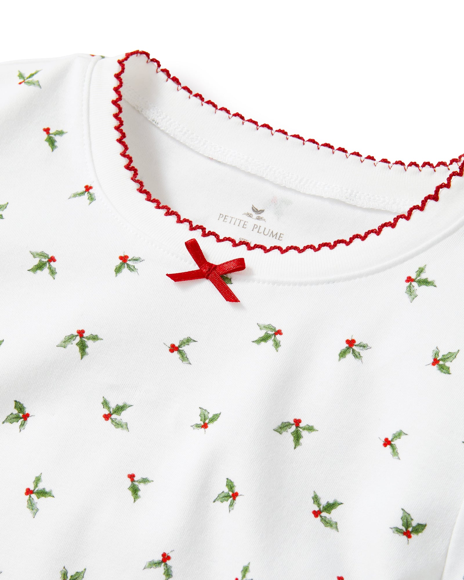 Kid's Pima Snug Fit Pajama Set in Mistletoe Moments: a two-piece white pajama set for children with a long-sleeve top, pants, green and red holly pattern, red scalloped neckline, and a red bow at the collar.